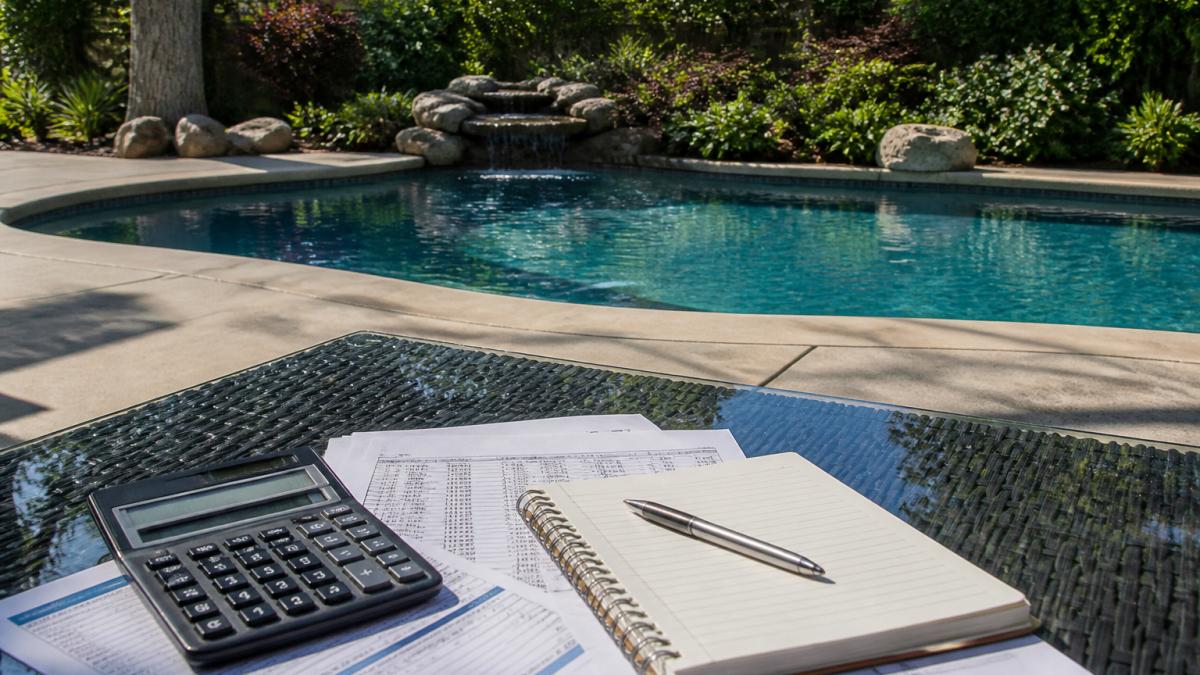 Completed residential inground pool with crystal-clear blue water, concrete decking surrounding the perimeter, and mature landscaping in a sunny backyard setting